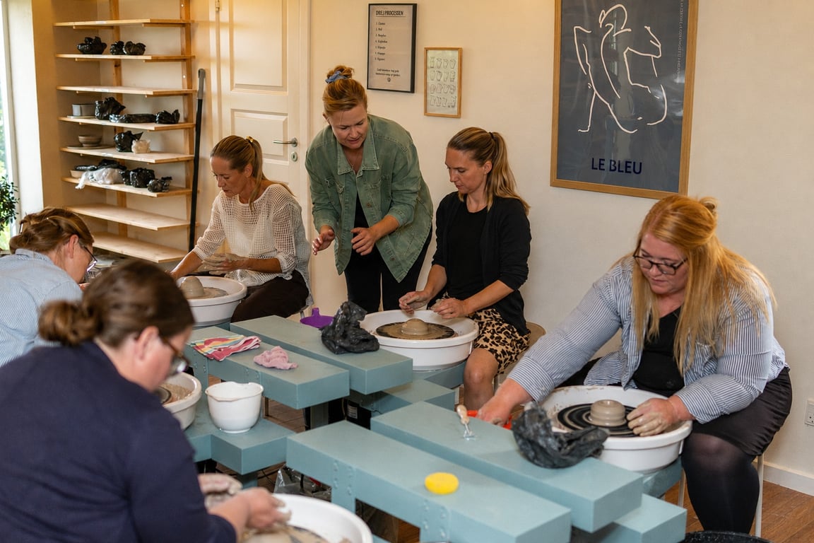 Gruppe af kvinder deltager i keramikworkshop, hvor de former ler på drejeskiver i et lyst, indendørs værksted.
