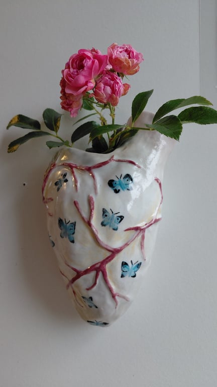 Keramisk hjerteformet vase med blå sommerfugle og røde årer, der indeholder pink roser og grønne blade, hængende på en væg.