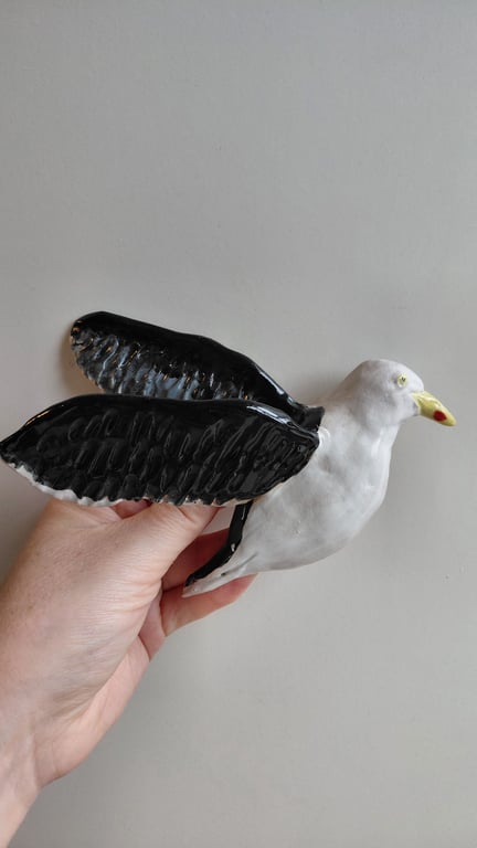Hånd, der holder en keramisk fugl med hvide krop og sorte vinger mod en grå baggrund.