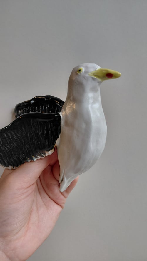 Hånd, der holder en keramisk fugl med hvid krop, sort vinge og gul næb med rød prik. (6)