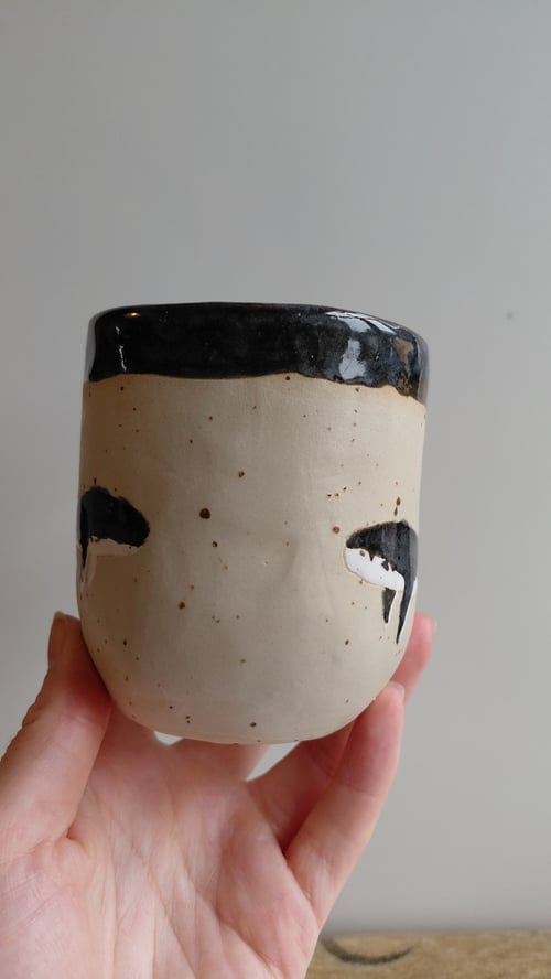 En hånd holder en lille keramikkop med sort glaseret kant og to sorte, malede øjenformer på en beige overflade. (3)
