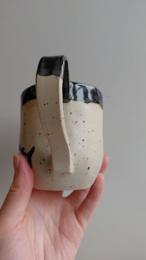 Hånd, der holder en beige keramik kop med sort glasur på kanten og hank. (7)
