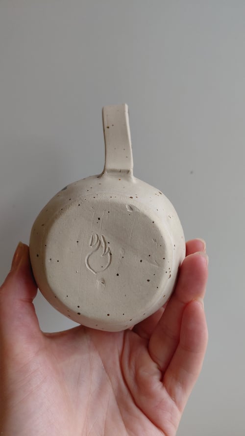 Hånd holder en lysegrå keramik kop set fra bunden med et brændemærke i form af en flamme. (9)