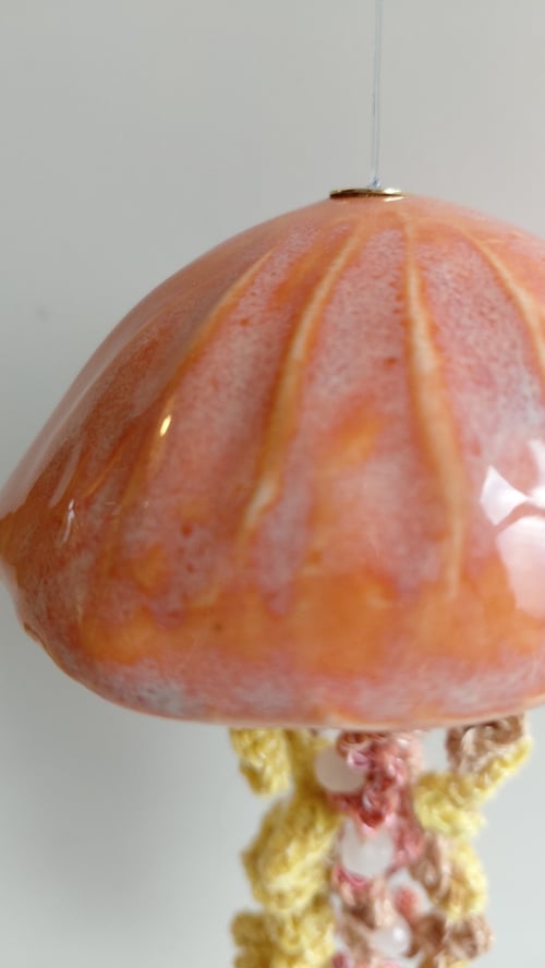 Nærbillede af en hængende, rosa-orange keramisk lampe med et mønster øverst og en hæklede dekoration i gule, rosa og brune nuancer under lampeskærmen. (6)