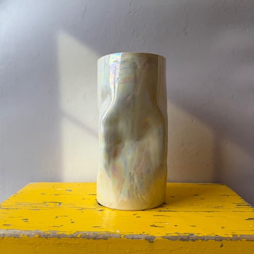 Glaseret keramisk vase med perlemorsglans placeret på et slidt, gult bord mod en hvid væg. (1)