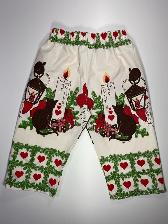 Bukser med julemotiv, inklusive sorte katte, tændte lys, lanterner, julegaver, gran og røde hjerter på hvid baggrund.
