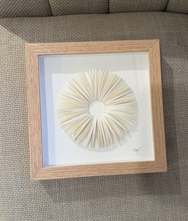 Billedramme med kunstværk bestående af foldede hvide papirsider arrangeret i en cirkel, placeret på en beige stofoverflade.