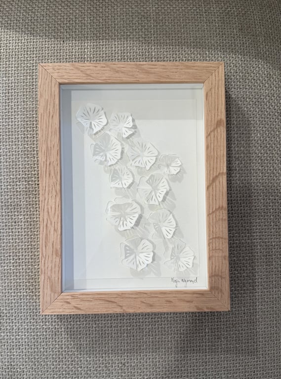 Indrammet papirkunst med flere hvide, udskårne og foldede blomster arrangeret diagonalt på en hvid baggrund, placeret på et groft vævet stofunderlag.