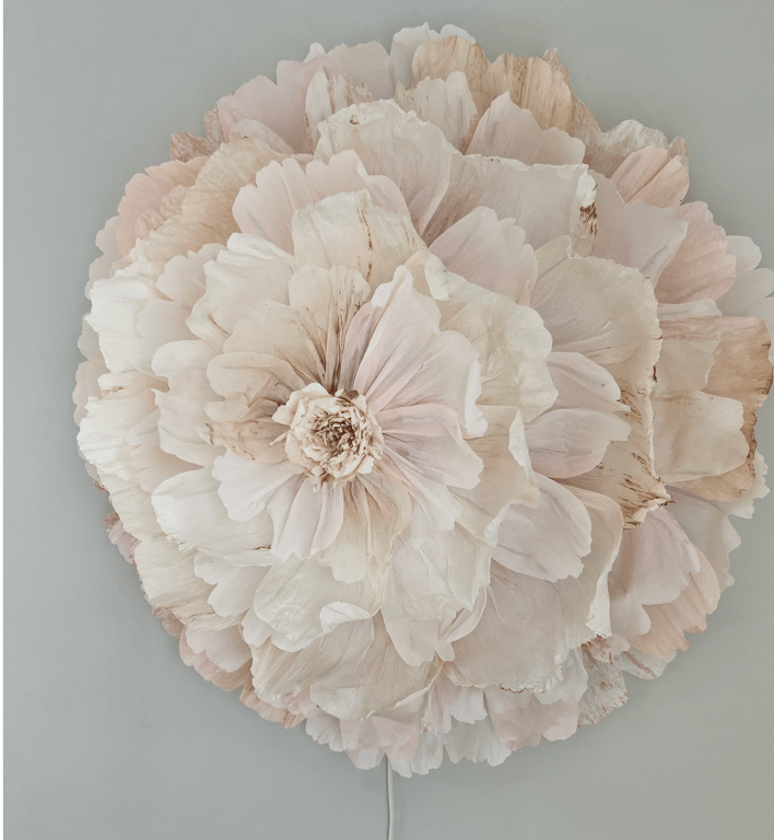 Stor, dekorativ blomst lavet af papir med lyserøde og beige nuancer, monteret på en grå væg.