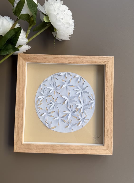 Træramme med cirkulært papirklip med blomsterdesign, placeret på en grå overflade ved siden af hvide blomster.