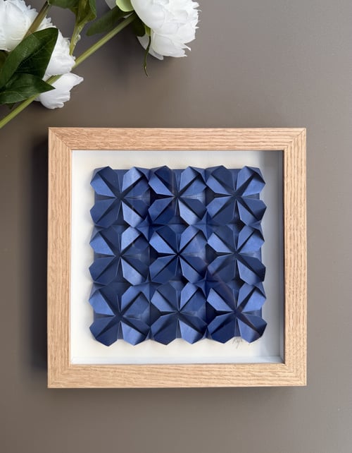 Indrammet papirkunst med tredimensionelle blå geometriske blomster på hvid baggrund, placeret på grå overflade med hvide blomster i øverste venstre hjørne. (1)