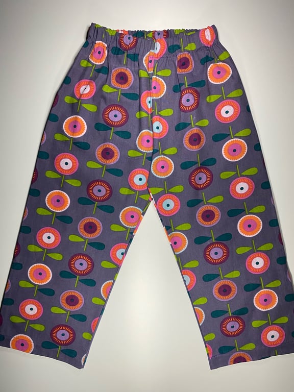 Børnebukser med elastisk talje i lilla stof med mønster af farverige, stiliserede blomster i rosa, pink, orange og grøn.