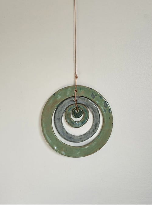 Hængende dekorativ ornament bestående af tre sammenkædede metalringe i forskellige størrelser med grøn og grå patina mod en lys væg. (1)