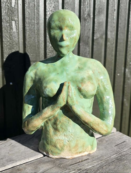 Skulptur af en menneskefigur i grønligt materiale med hænderne samlet i bøn foran brystet, placeret på et træbord med en mørk baggrund. (1)