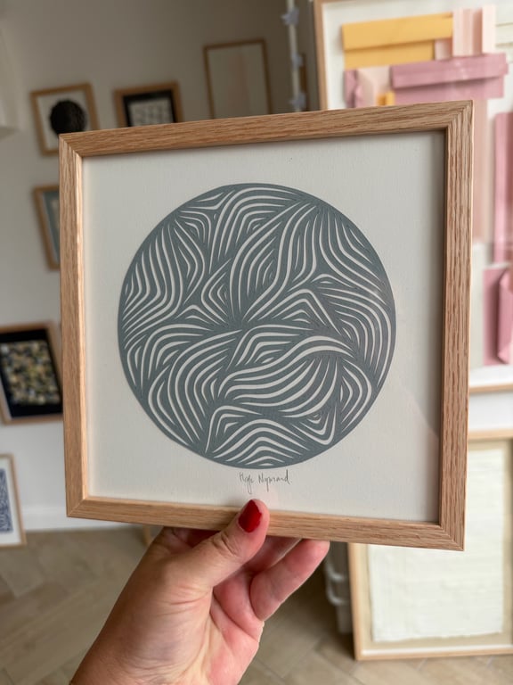 Hånd, der holder en indrammet rund abstrakt kunstplakat med bølgende, linjemønstre i gråtoner.