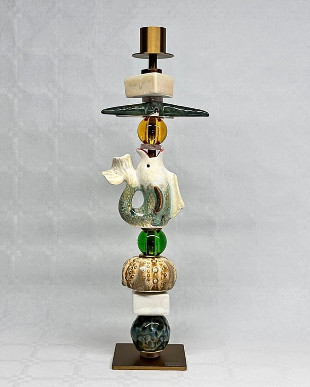 Skulptur bestående af forskellige keramiske og glas-dele stablet lodret, herunder en fisk, kugler og geometriske former, på en metalplade mod grå baggrund.