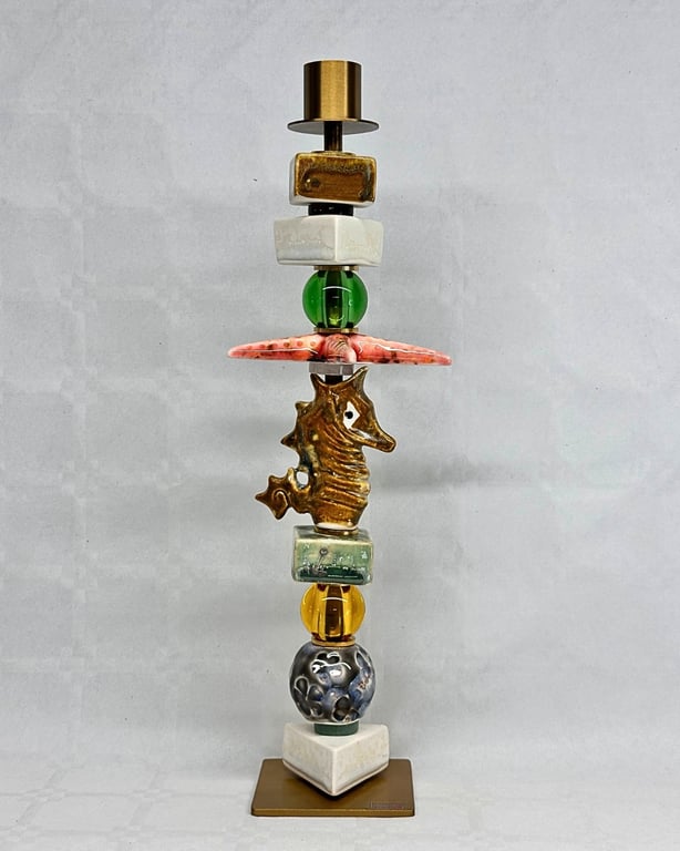 Farverig skulptur bestående af stablede kunstneriske perler og figurer, inklusive en gylden søhest, på en brun firkantet base mod grå baggrund.
