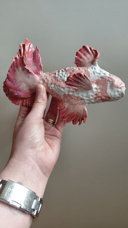 Hånd, der holder en skulptur af en fisk med røde og hvide nuancer mod en grå baggrund.