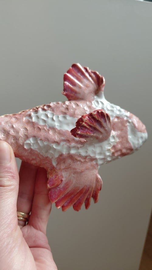 Hånd holder en keramisk fiskefigur med mønster i hvide og røde nuancer mod en grå baggrund. (3)