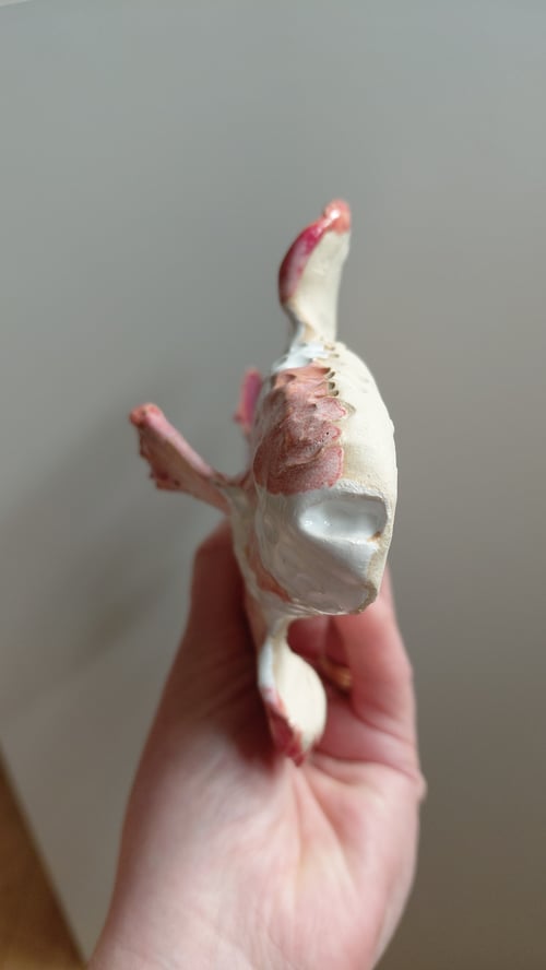 Hånd, der holder en delvist malet og beskadiget hvid og pink plastik- eller keramisk figur set fra undersiden. (7)