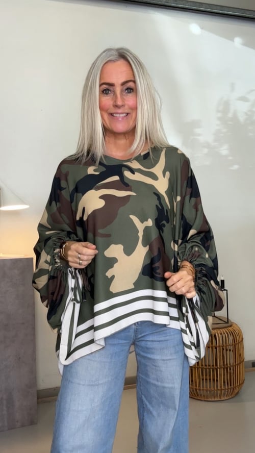 Kvinde med langt blondt hår iført en camouflage-mønstret bluse med løse ærmer og lyseblå jeans, stående i et lyst rum med moderne indretning. (1)