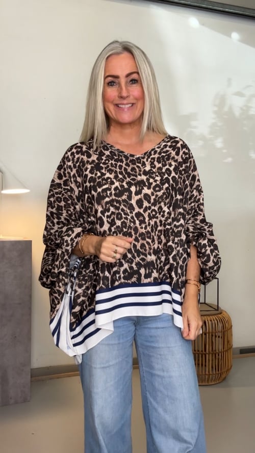 Kvinde med langt blond hår, iført leopardmønstret bluse med stribede kanter og lyseblå jeans, stående indendørs foran væg. (1)