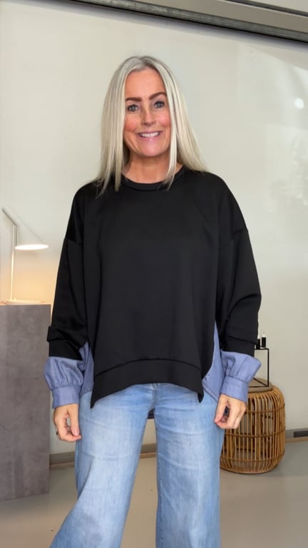 Kvinde med langt blondt hår iført sort og blå bluse og lyseblå jeans, stående indendørs foran hvid væg.