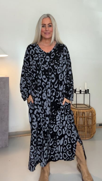 Kvinde med langt blond hår iført lang, løs kjole med sort og gråt leopardmønster og beige støvler, stående i lyse rum med kurvestol og stearinlys på baggrund.