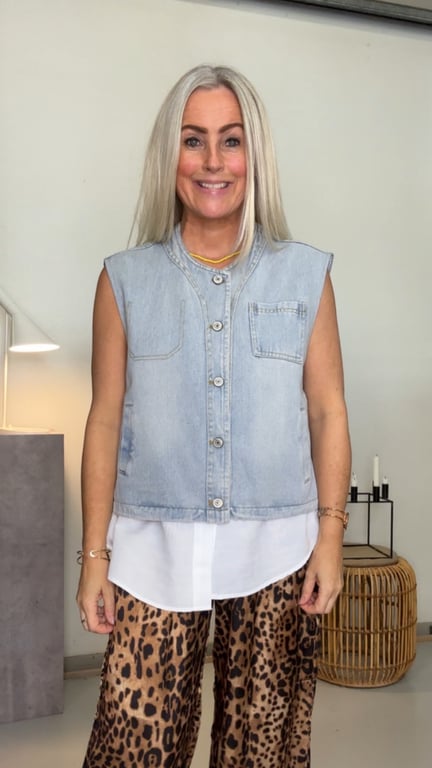 Kvinde med langt lyst hår iført lyseblå denimvest, hvid skjorte og leopardmønstrede bukser, stående i et lyst rum med møbler og dekorative genstande.