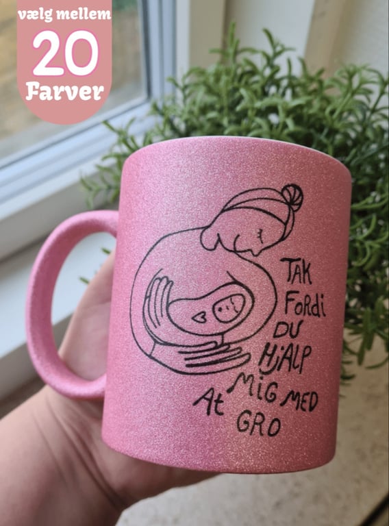 Lyserød glitrende krus med tegning af en person, der holder en baby, og teksten "TAK Fordi DU HjALP MIG med At GRO", holdt i en hånd foran en vindueskarm med grøn plante. Tekst øverst til venstre siger "vælg mellem 20 Farver".