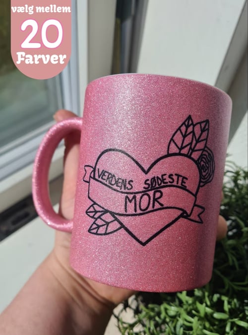 En hånd holder en glitrende lyserød krus med sort hjerte og tekst "Verdens sødeste mor" foran en vindueskarm med en grøn plante. I øverste venstre hjørne er der en tekstboble med teksten "vælg mellem 20 farver". (1)