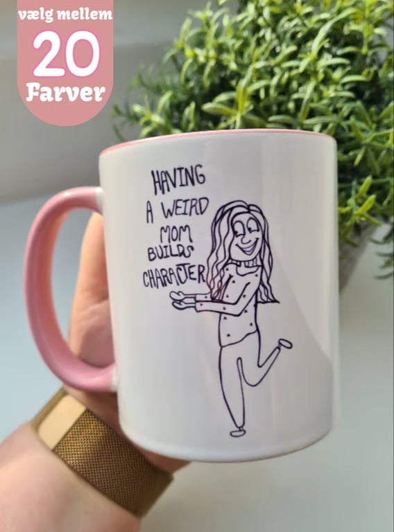 En hånd holder en hvid krus med lyserød hank og kant, med tekst og tegning af en glad kvinde, der siger "HAVING A WEIRD MOM BUILDS CHARACTER". I baggrunden er grønne planter, og et skilt øverst til venstre viser "vælg mellem 20 Farver".