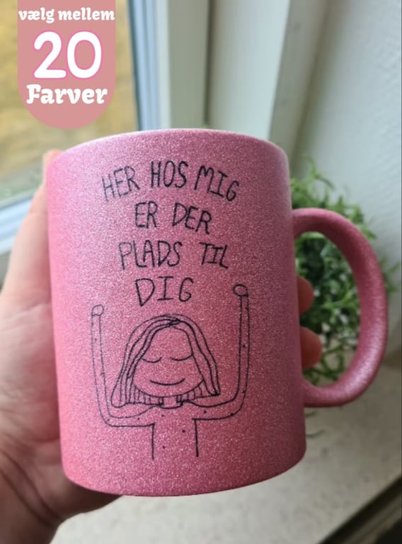 En hånd holder en pink glitrende krus med tegning af en afslappet person og teksten "Her hos mig er der plads til dig". En tekst i øverste venstre hjørne siger "vælg mellem 20 farver".