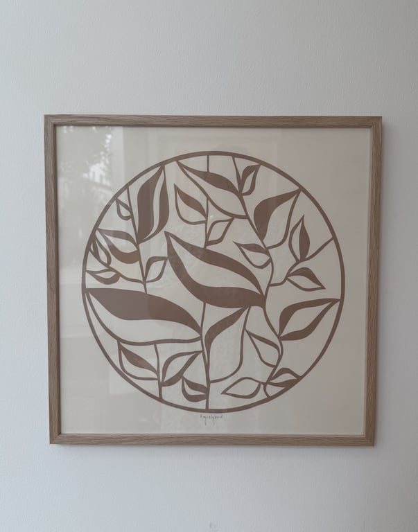 Billede af en firkantet, træramme med en kunstplakat indeholdende en cirkel fuld af stiliserede blade i brunlige nuancer mod en beige baggrund.