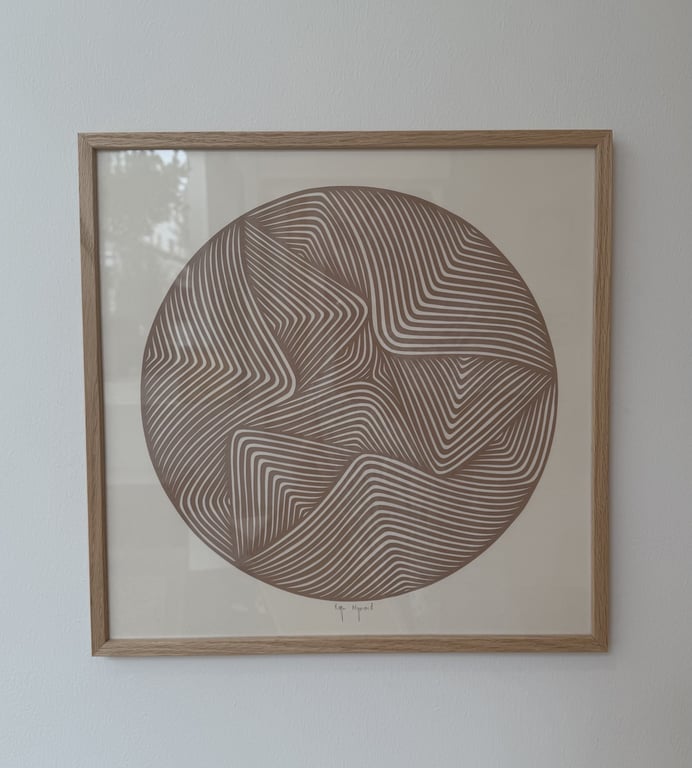 Abstrakt kunstplakat med en cirkel fyldt med brune og hvide geometriske linjemønstre, indrammet i en enkel lys træramme og hængt på en hvid væg.