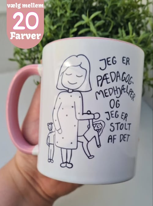 Hånd holder hvidt krus med pink hank og tegning af pædagog med børn samt teksten "Jeg er pædagogmedhjælper og jeg er stolt af det". Tekst på billedet: "vælg mellem 20 farver". (1)