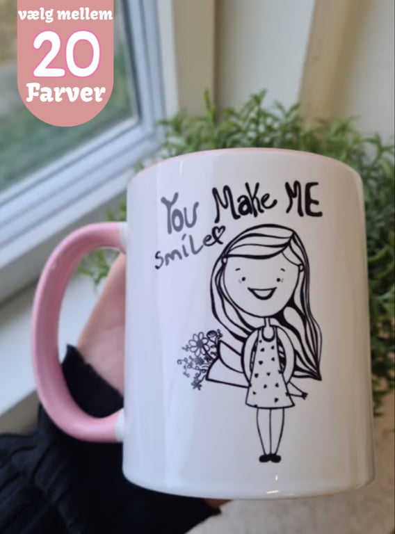 Hånd der holder en hvid krus med lyserødt håndtag og inderside, dekoreret med en tegning af en smilende pige med en buket blomster og teksten "You Make Me Smile". Tekst i øverste venstre hjørne på dansk: "vælg mellem 20 farver".