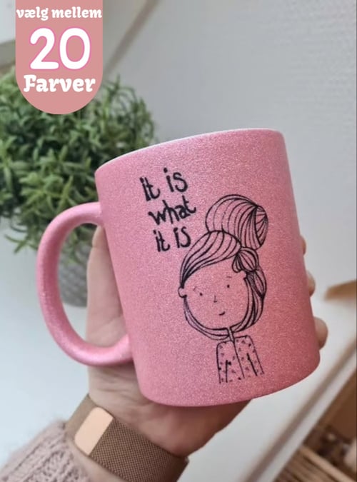 Hånd holder pink glitrende krus med tegning af pige og teksten "It is what it is", samt tekst øverst "vælg mellem 20 farver". (1)