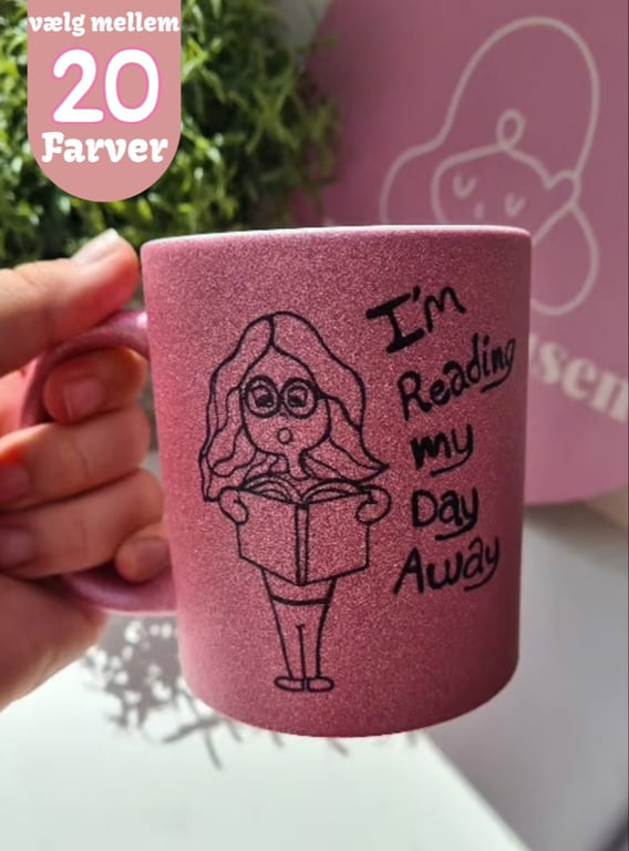 En hånd holder et glitrende lyserødt krus med en tegning af en person med briller, der læser en bog, og teksten "I'm Reading my Day Away". I øverste venstre hjørne er teksten "vælg mellem 20 Farver".