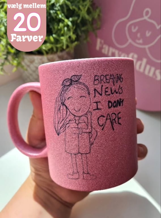 Lyserød glitrende krus med tegning af smilende pige og teksten "BREAKING NEWS I DON'T CARE", holdt i en hånd. Tekst i øverste venstre hjørne: "vælg mellem 20 farver".