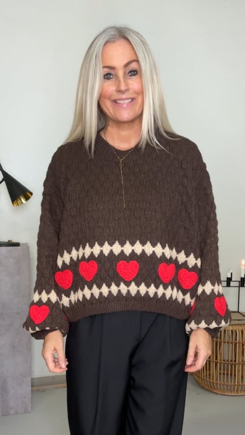 Kvinde iført brun, strikket sweater med røde hjerter og sort bukser, stående indendørs foran hvid væg. (1)