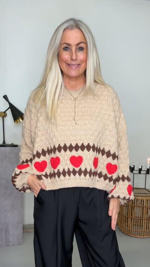 Kvinde med langt blond hår iført beige striktrøje med røde hjerter og sort buks, stående foran hvid væg med lampe og dekorative lys. (1)