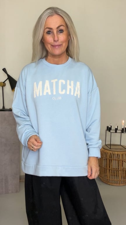 Kvinde med langt blond hår iført lyseblå sweatshirt med teksten "MATCHA CLUB" og sorte bukser, stående i et lyst rum med moderne indretning.