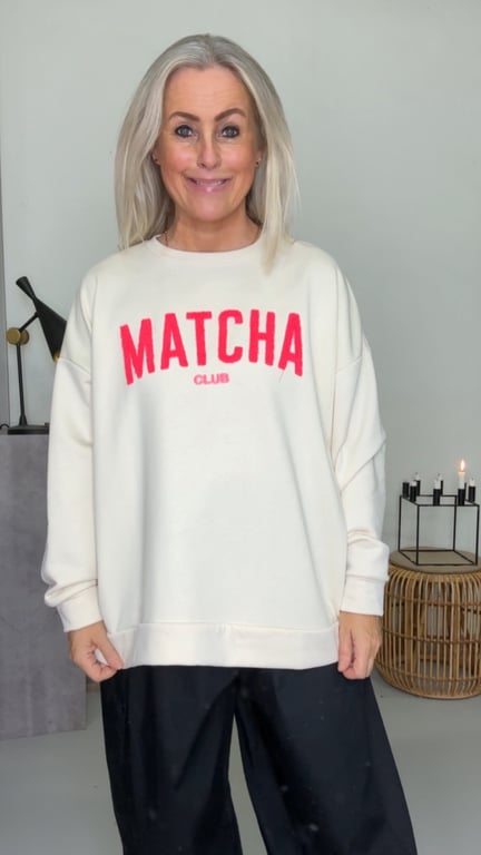 Kvinde med langt gråt hår iført hvid sweatshirt med teksten "MATCHA CLUB" i rødt og sort bukser, står foran en lys væg med dekorative elementer som stearinlys og en lampe.