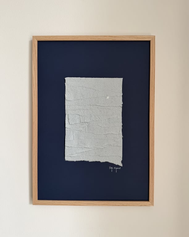 Billede af en kunstplakat med en hvid, tekstureret papirfigur på en mørkeblå baggrund, indrammet i en lys træramme.