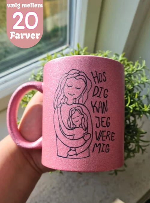 Hånd, der holder en rosa glitrende krus med illustration af to personer, en der omfavner en anden, og teksten "Hos dig kan jeg være mig". Tekst øverst til venstre: "vælg mellem 20 farver". (1)