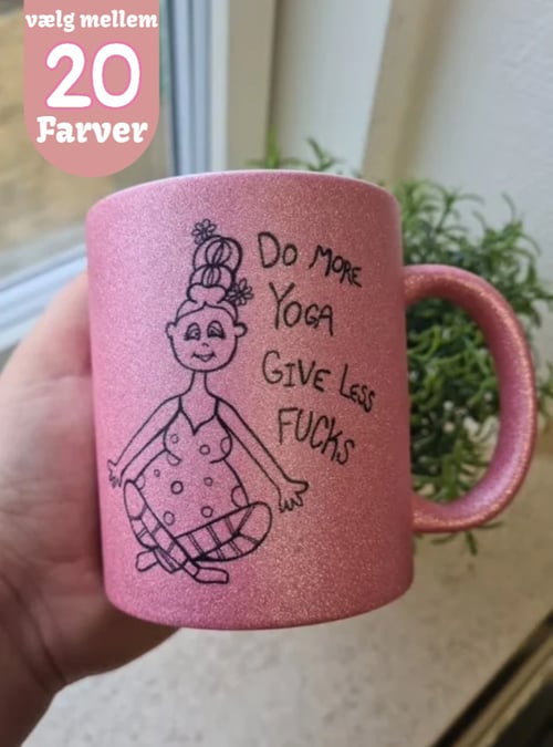 Hånd holder en pink glitrende kop med tegning af en kvinde i yogastilling og teksten "Do more yoga give less fucks". Tekst i hjørnet: "vælg mellem 20 farver". (1)