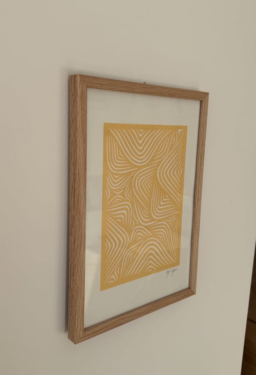 Billede af en væg med en indrammet gul kunstplakat, der har hvidt, abstrakt mønster i bølgende linjer, i en træramme. (1)