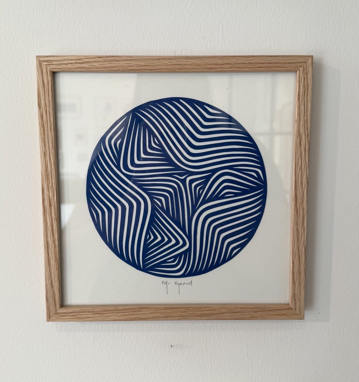 Indrammet kunstprint af en blå cirkel med hvidt geometrisk mønster bestående af buede linjer, hængende på en hvid væg.