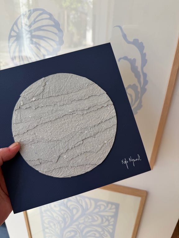 Hånd, der holder et kunstkort med en rund tekstureret grå cirkel på mørkeblåt papir, underskrevet af kunstneren.